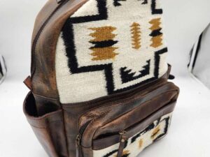 A brown leather backpack with Southwestern-style fabric panels featuring geometric patterns in white, black, and tan. The design includes a main compartment and two front pockets. The backpack is displayed against a plain background.