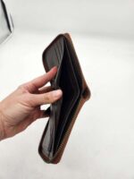 A hand holding an open brown leather wallet with a zipper. The wallet appears mostly empty, with visible card slots and compartments inside. The background is plain white, emphasizing the wallet and the hand.