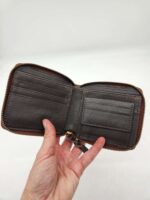 A hand holding an open brown leather wallet with a zipper closure. The wallet has multiple card slots on the left and an empty pocket on the right. The background is plain and white.