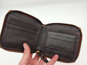 A hand holding an open brown leather wallet with a zipper around the edge. The wallet has multiple card slots on the left side and a smaller pocket on the right side. The background is plain white.