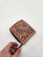 A hand is holding a small, intricately designed leather wallet with a brown embossed floral and feather pattern. The wallet features a zipper closure and is placed on a white background.