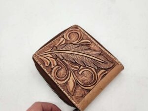 A hand is holding a small, intricately designed leather wallet with a brown embossed floral and feather pattern. The wallet features a zipper closure and is placed on a white background.