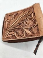 A close-up shot of a brown leather wallet featuring intricate embossed designs. The decorative pattern includes a prominent feather amidst swirling, leaf-like shapes. The wallet has a zipper closure with a braided leather pull tab.