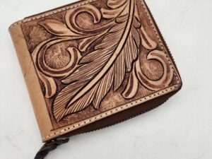 A brown leather wallet with an ornate, embossed leaf and swirl design on the front. The wallet has a zipper closure with a small, braided leather pull tab. The background is a plain, white surface.