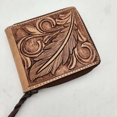 A brown leather wallet with an ornate, embossed leaf and swirl design on the front. The wallet has a zipper closure with a small, braided leather pull tab. The background is a plain, white surface.