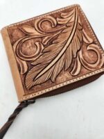 A brown leather wallet with intricate carvings of a large feather and swirling patterns on the front. The wallet has a braided leather zipper pull and sits on a white surface.