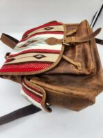 A brown leather backpack with a flap featuring a colorful woven fabric design consisting of red, white, and blue patterns. The flap is secured with a brass buckle. The bag has dark brown straps and a drawstring closure. The image is set against a plain white background.