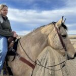 A woman in a green sweater and blue jeans rides a light brown horse with a Red Buckstitch- Breast Collar on a dirt path, surrounded by wide-open fields and under a partly cloudy sky.