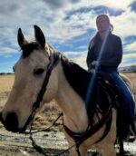 A person with blonde hair rides a light-colored horse along a dirt path, the horse sporting a Red Buckstitch- Breast Collar. The sky above is partly cloudy with the sun shining brightly, creating a sunbeam effect in the image. They are dressed in a dark jacket and jeans, with open fields in the background.