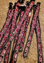 A pile of decorative western-style belts with intricate red, black, and white patterns is laid out on a brown carpet. The belts have silver buckles and price tags attached.