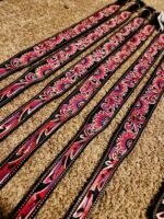 A row of intricately designed leather belts featuring vibrant red and pink patterns with black and white accents displayed on a beige carpeted surface. Each belt has a unique but cohesive detailed decorative motif.