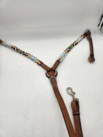 A horse breast collar with a pattern of multicolored beads on white straps, brown leather accents, and silver hardware, is laid flat on a white surface. One end has a clip and the other ends loop for attachment.