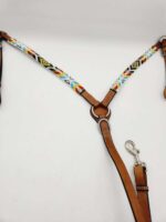 A leather harness with colorful woven fabric adornments. The fabric features geometric patterns in red, yellow, blue, and black on a white background. The harness has adjustable straps, a metal ring, and a metal clasp for attaching a leash. It is displayed on a white background.