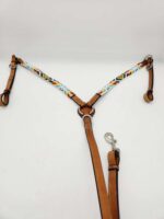 A tan leather dog harness with a colorful, patterned strap across the chest. The harness features various buckles, rings, and an attached leash with a metal clip. The background is a plain white surface.