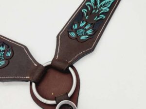 Close-up of a brown leather harness with teal floral embroidery and silver hardware. The harness features a circular metal ring at the center where the leather pieces connect and a silver clip attached to one end. The background is white.