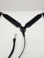 A black leather harness with silver hardware is laid out against a plain white background. The harness features intricate stitching and has a central metal ring connecting the straps and the leash.