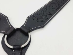 A close-up image of a black leather harness with engraved floral designs. It features a silver metal ring connecting two leather straps, which are stitched with white thread along the edges. The background is plain white.