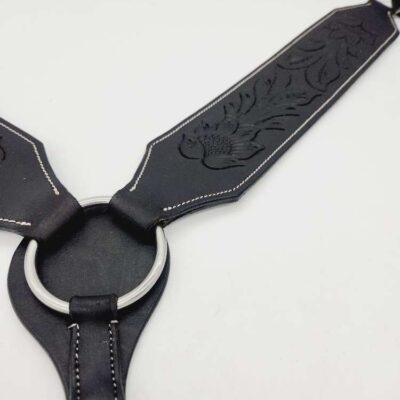 A close-up image of a black leather harness with engraved floral designs. It features a silver metal ring connecting two leather straps, which are stitched with white thread along the edges. The background is plain white.