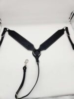 A black leather harness with intricate patterns, featuring metal rings and clips, is laid out on a white surface. The harness is structured with two broad, decorative leather straps connected by a circular metal ring, and includes an attached leash.