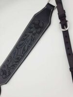 A close-up of a black leather guitar strap with intricate floral and feather embossing. The strap has a silver buckle and adjustable holes for fitting. The detailed craftsmanship highlights the texture and pattern on the leather.