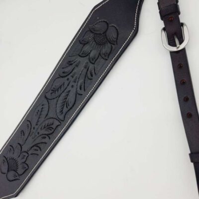 A close-up of a black leather guitar strap with intricate floral and feather embossing. The strap has a silver buckle and adjustable holes for fitting. The detailed craftsmanship highlights the texture and pattern on the leather.
