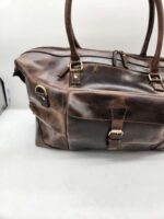 A distressed brown Leather Duffel Bag- City Square with two top handles, a front pocket secured with a buckle, and a zippered main compartment. The view shows the front and part of the side of the bag, highlighting its rustic texture and hardware details. Includes a convenient shoe pocket.