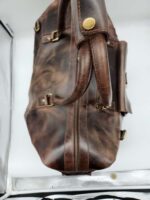 A Leather Duffel Bag- City Square with brass hardware and a zipper runs along the side. The bag has a worn, vintage appearance and features a handle at the top. It also includes a convenient shoe pocket. The photo is taken in a light-colored setting.