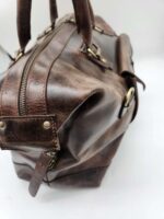 A close-up image of a Leather Duffel Bag- City Square with visible handles and a large zipper on the top. The bag features brass hardware, including buckles and rivets, with a slightly worn but sturdy appearance. Complete with a convenient shoe pocket, the background is plain white.