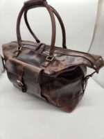 A Leather Duffel Bag- City Square with two sturdy handles and a large front pocket secured by a buckle. The bag features a full-length zipper on top, metal hardware accents, and an additional shoe pocket for extra convenience. The backdrop is plain and light, highlighting the rugged texture of the leather.