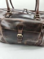 A close-up view of a Leather Duffel Bag- City Square with a buckle clasp on the front pocket and two leather handles. The bag shows some worn and distressed detailing, adding to its vintage appearance. A zipper is visible on the top of the bag. The background is plain white.