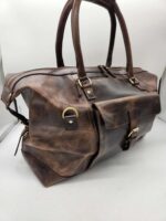 A Leather Duffel Bag- City Square with a worn vintage look, featuring a dedicated shoe pocket. It has sturdy handles, brass hardware, and multiple pockets and buckles for secure closure and convenience. The bag is placed against a plain white background.