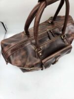 A brown Leather Duffel Bag- City Square with multiple zippers and handles is placed on a white surface. The bag exudes a rugged, vintage appearance with brass hardware accents, an exterior pocket, and a dedicated shoe pocket for added convenience.