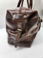 A close-up image of a Leather Duffel Bag- City Square, showcasing its textured surface and detailed stitching. The bag has a rugged look with a zipper on top, a strap with brass hardware, and features an integrated shoe pocket. The image highlights the craftsmanship and quality of the leather material.