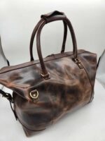 A weathered brown Leather Duffel Bag- City Square with dual handles and brass hardware, including buckles and D-rings, is shown against a plain white background. Featuring a convenient shoe pocket, the bag has a worn, rugged appearance with visible creases and patina.