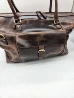 A well-worn, dark brown leather tote bag with sturdy handles is shown. The bag features a large front pocket secured with a buckle strap and zipper, and has metal hardware accents. With a vintage look from its slightly distressed surface, this Leather Duffel Bag- City Square also includes a convenient shoe pocket.