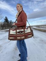 A person with long hair draped over one shoulder stands outside on a snowy path, holding a large, patterned shoulder bag. They are wearing a rust-colored jacket, blue jeans, and boots. A cloudy sky and a wooden fence are visible in the background.