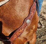 Close-up of a brown horse wearing an ornate leather chest strap with intricate swirl patterns and a decorative silver concho. A person in blue jeans and brown boots is partially visible in the background, standing beside the horse on a dirt surface.