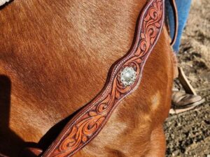 Close-up of a brown horse wearing an ornate leather chest strap with intricate swirl patterns and a decorative silver concho. A person in blue jeans and brown boots is partially visible in the background, standing beside the horse on a dirt surface.