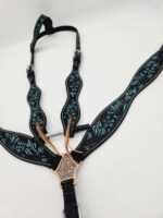 A detailed close-up of an ornate horse breast collar lies on a plain white surface. The black leather collar is embellished with intricate turquoise floral patterns and features engraved metallic accents at the junctions.