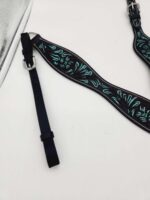 Close-up of a black leather strap featuring an embossed, intricate turquoise floral design. The strap has a shiny metal buckle and an adjustable portion with multiple holes. It is laid out on a white surface, with a part of a mirror visible in the top left corner.