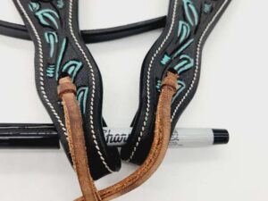 A close-up of intricately designed leather horse tack with blue and black floral patterns. The tack features brown leather straps and is positioned on top of a black and silver Sharpie marker, which is partially visible beneath the straps.