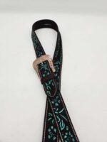 A stylish black leather belt with embroidered turquoise-blue patterns is coiled on a white surface. The belt features a decorative and intricate silver buckle. The detailed embroidery contrasts sharply with the smooth black leather.