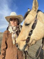 A person wearing a brown jacket, patterned scarf, and cowboy hat stands outdoors beside a light brown horse. The sky is clear and blue, suggesting a sunny day.