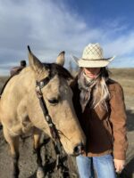 A woman wearing a cowboy hat, brown jacket, and blue jeans stands close to a tan horse with a bridle. They are outdoors under a blue sky with some clouds, on a dirt surface. The woman and horse appear calm and relaxed.