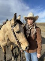 A woman wearing a cowboy hat and brown jacket stands beside a horse. The horse has a light tan coat and a bridle on its head. The background shows an open landscape with a blue sky and some clouds.