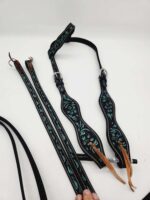 A black and teal western browband headstall is displayed against a white background. The headstall features intricate teal patterns and silver buckles, with leather ties at the ends. The reins, adorned with matching teal designs, are lying beside it.