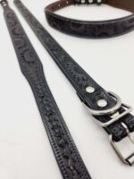 A close-up of two black leather dog collars lying on a white surface. The collars feature intricate embossed floral patterns and have silver buckles and hardware. One collar is positioned straight, while the other is coiled in the background.