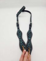 A decorated leather guitar strap with a black base and intricate teal patterns is laid out on a white surface. The strap features two adjustable buckles and two leather string ends. A hand is holding the string ends.