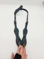 A black guitar strap with teal decorative patterns is laid on a white surface. It features two adjustable buckles and a pair of brown leather ties. A hand wearing a black wrist strap is holding the end of the strap with the ties.