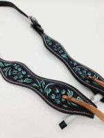 A close-up of a black leather guitar strap featuring intricate turquoise floral and leaf designs. The strap is adorned with a brown suede lace and has a small adjustment buckle. A white marker with a black cap is tucked under the strap for scale.